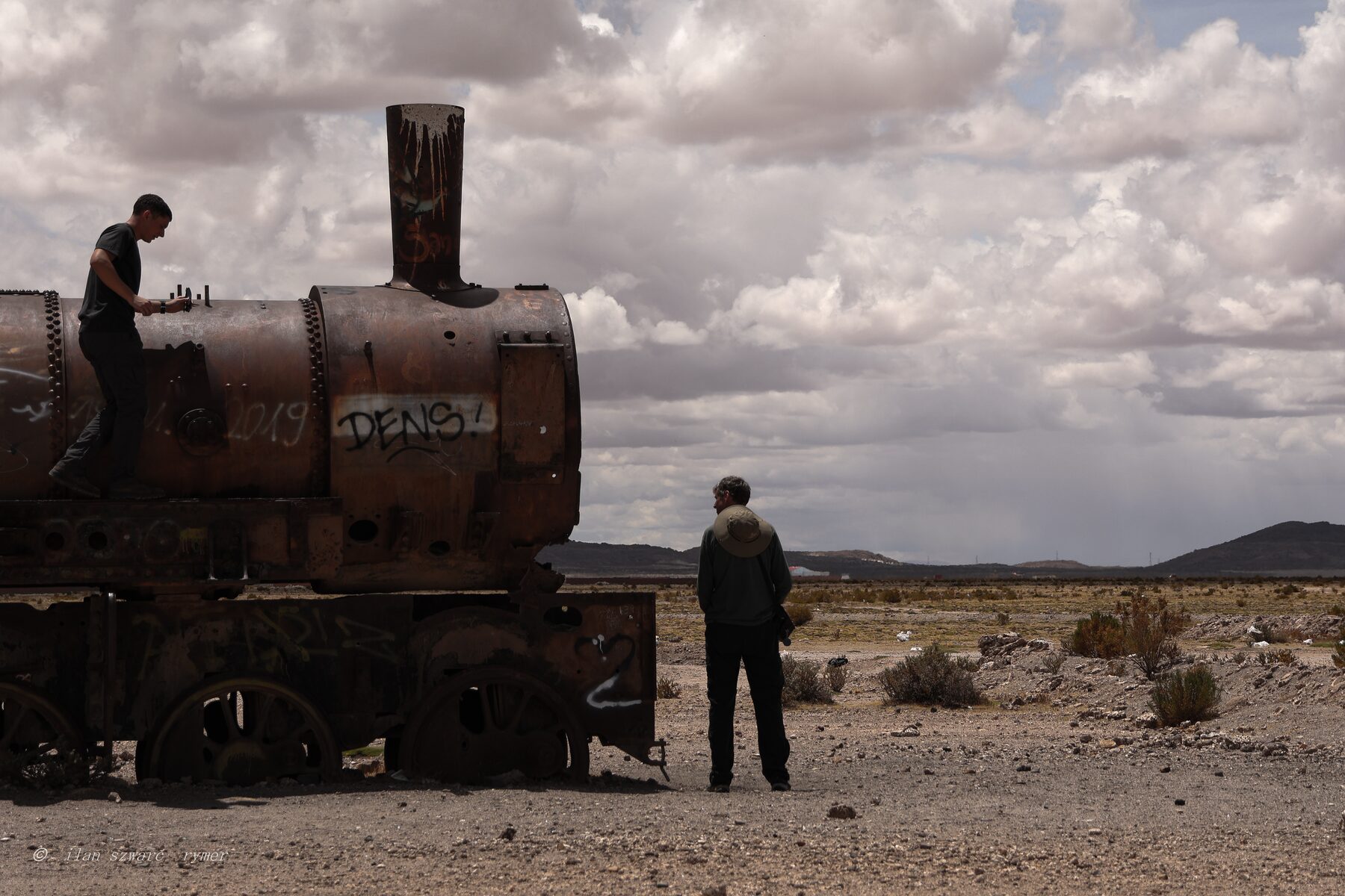 Two figures beside a rusted steam locomotive in a wide desert landscape